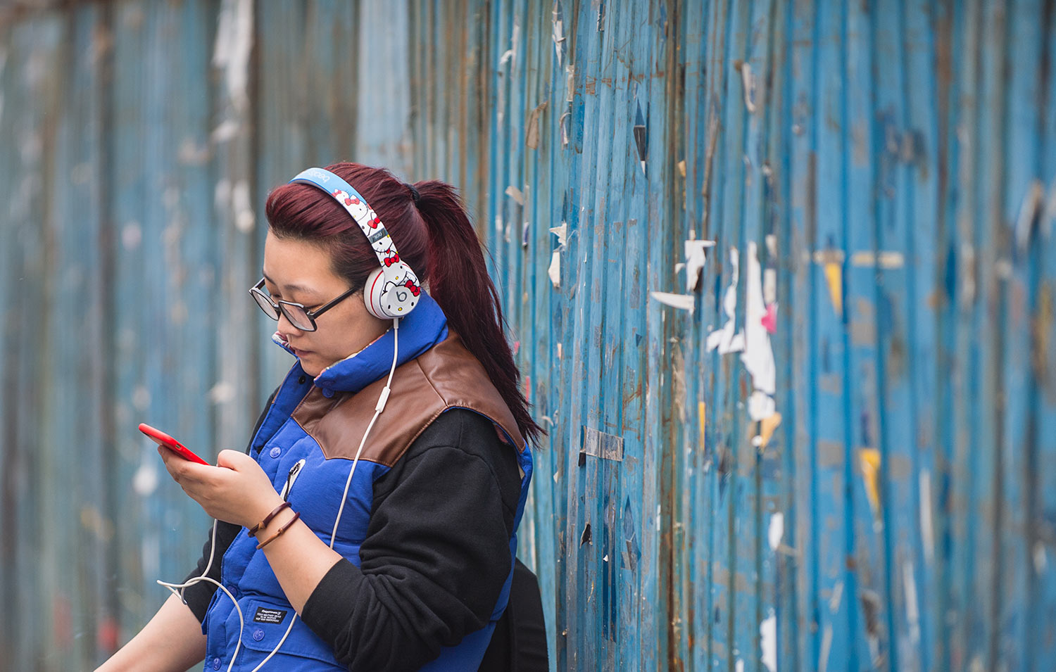 A Chinese woman wearing Hello Kitty headphones looks at her phone while she stands beside a blue fence.