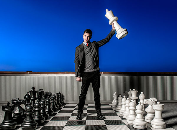Derek Pacqué, the founder of CoatChex, stands on a rooftop in Long Island City with larger-than-life chess pieces. The Manhattan skyline is in the distance.